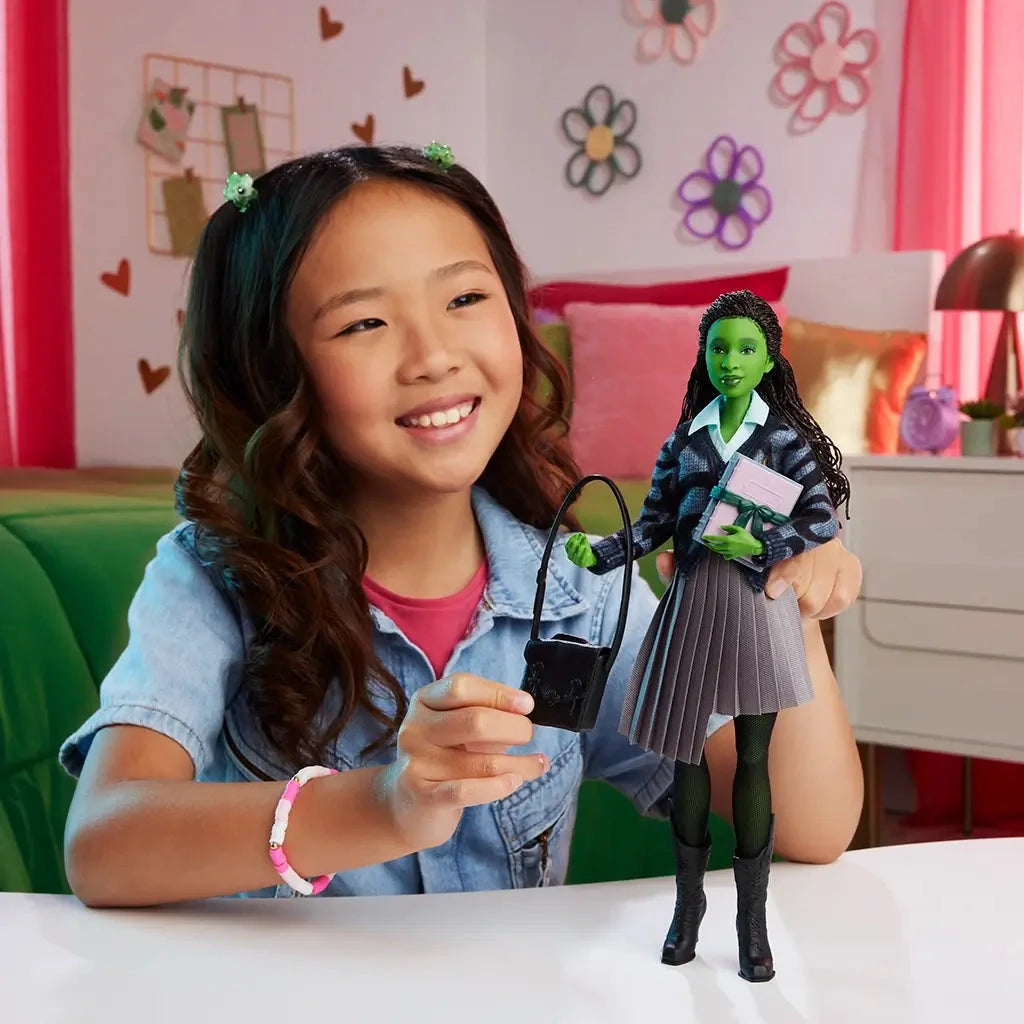 Young girl smiling while holding Wicked Elphaba doll, displaying the black bag and pink book accessories in a colorful bedroom setting.