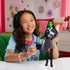 Young girl smiling while holding Wicked Elphaba doll, displaying the black bag and pink book accessories in a colorful bedroom setting.
