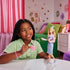 Young girl smiling while playing with Wicked Glinda doll, holding pink purse accessory in a colorful bedroom setting.