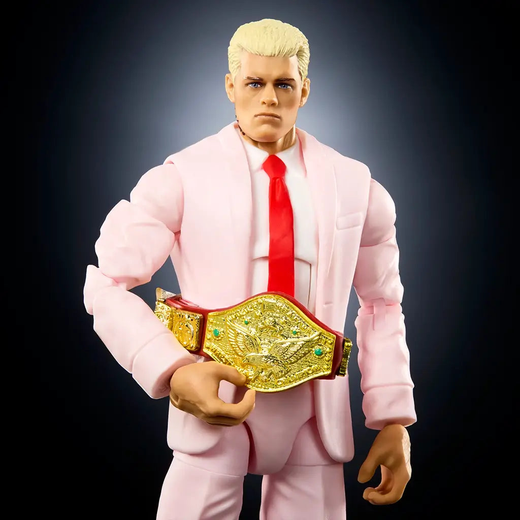WWE Cody Rhodes action figure close-up showing light pink suit fabric sculpting, smooth plastic finish, blond hair, blue eyes, neutral facial expression, red tie detail, and championship belt accessory held across the torso against a dark studio background.