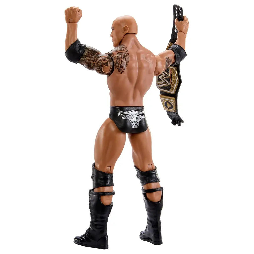 Rear angled view of The Rock WWE action figure reveals sculpted tribal tattoos, black boots, raised fists, and gold-trimmed championship belt details displayed in a bright white studio setting.