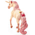 Schleich bayala decorated unicorn mare has a pale cream coat, shimmering pink mane and curled tail with red jeweled bands, shown from behind with a golden horn on a white background.